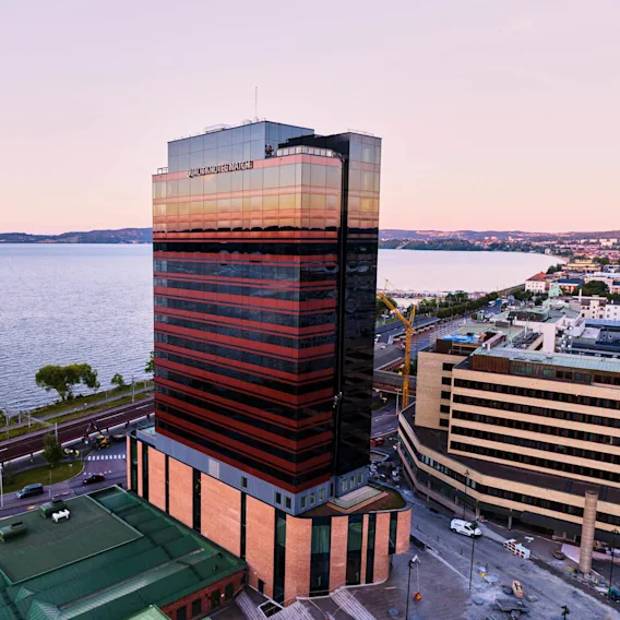 En høy, speilvendt bygning med teksten "QUICK OFFICE" reflekterer en solnedgang over en stor innsjø, plassert i et urbant miljø med veier og tilstøtende strukturer.