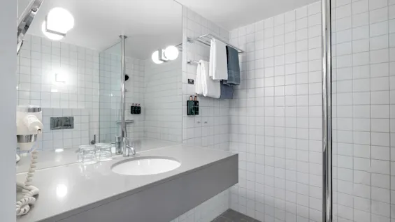 Et rent, moderne hotellbad med vask, speil, hårføner og dusjområde med friske håndklær.