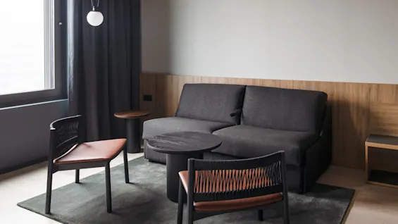 A gray sofa sits in a minimalist, modern living room with a small round table and two chairs on a rug. A pendant light hangs near a curtained window.