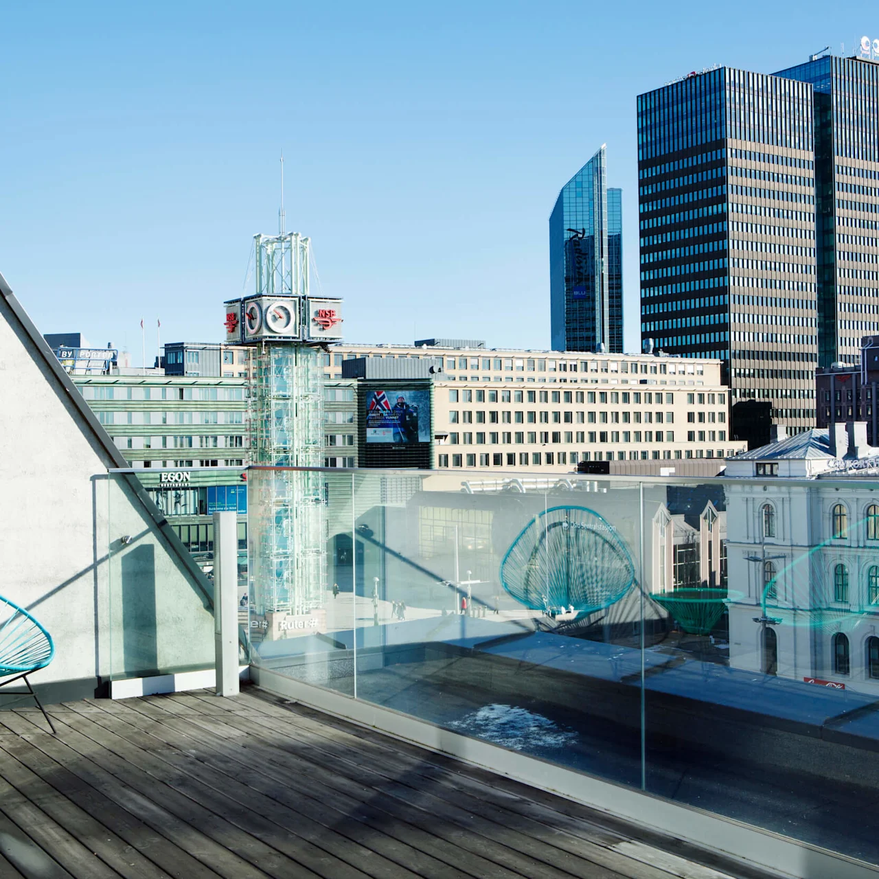Two turquoise chairs sit on a wooden rooftop terrace overlooking modern city buildings, including a clock tower. Sunlight casts shadows, and glass railings provide an unobstructed view.