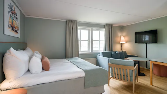 En pent redt seng med puter og teppe, ved siden av et lite bord og stol. En sofa og lampe er i bakgrunnen nær et vindu med gardiner i et koselig, mykt opplyst rom.