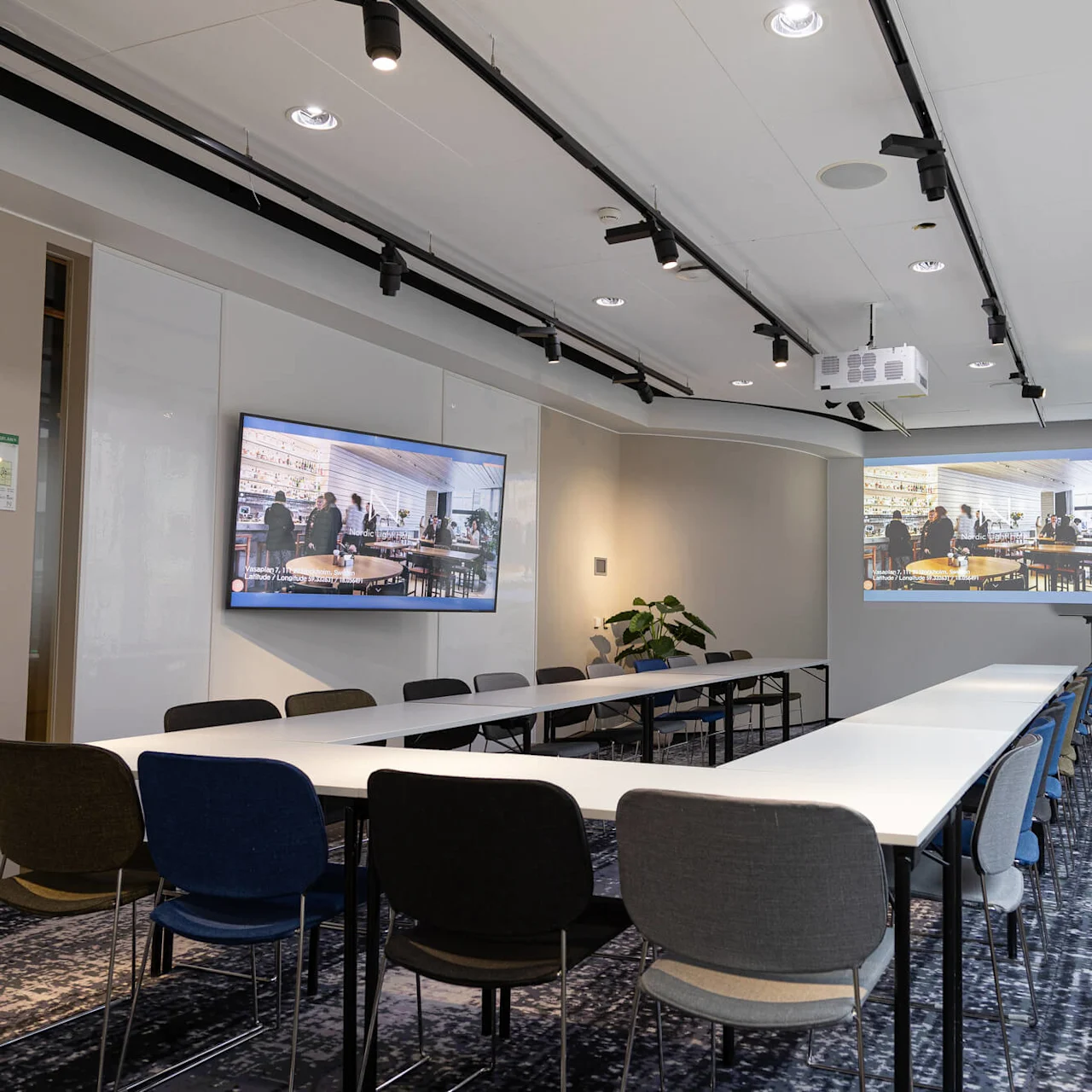 Conference room with tables and chairs arranged in a U-shape. Two screens display an image of a busy restaurant. Ceiling features spotlights, walls are plain, and windows line the side.