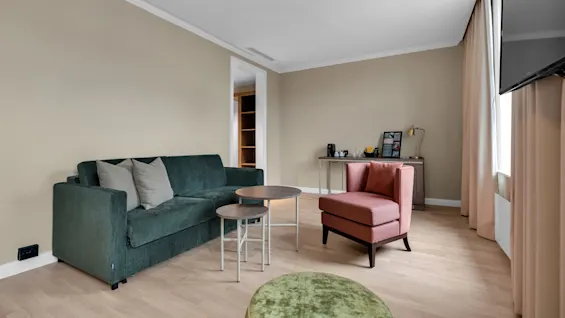 Ett snyggt vardagsrum med en grön soffa, rosa fåtölj, hoplagda soffbord och en TV på väggen, i naturligt ljus.