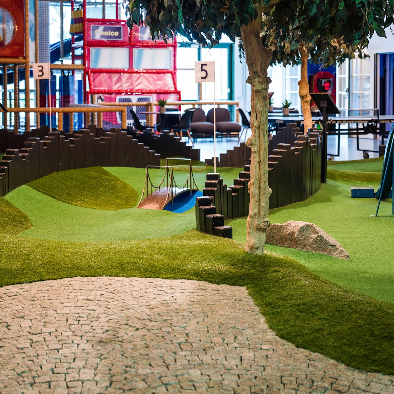 A mini-golf course sits indoors with numbered holes, artificial grass, and a tree, bordered by a stone path. In the background, a colorful play area for children is visible.