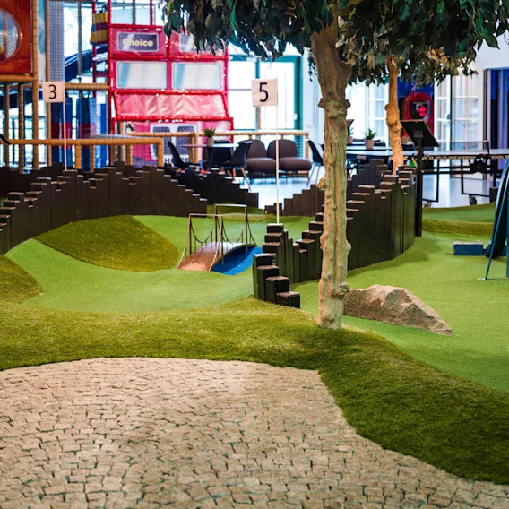 En minigolfbane er indendørs med nummererede huller, kunstgræs og et træ, omgivet af en stensti. I baggrunden ses et farverigt legeområde for børn.