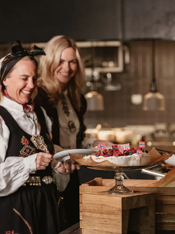 To kvinner i bunad smiler ved et buffetbord med en pavlova pyntet med norske flagg.