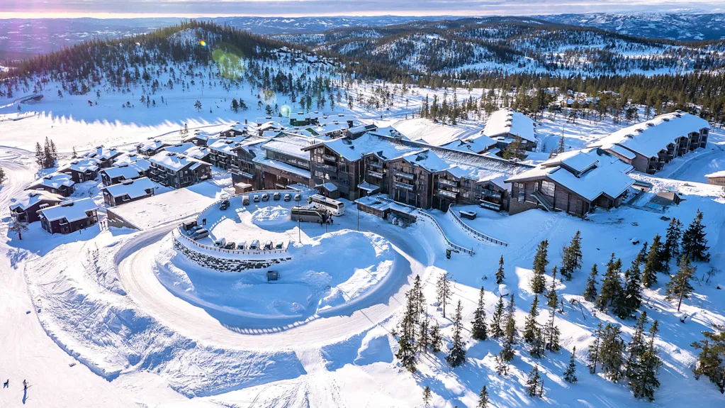 Suuri, luminen lomakeskuskokonaisuus sijaitsee vuoristoisessa maisemassa mäntyjen keskellä, ympärillään kiemurtelevat lumiset polut ja pysäköidyt ajoneuvot.
