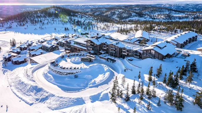 A large, snow-covered resort complex is nestled in a mountainous landscape with pine trees, surrounded by winding snowy paths and parked vehicles. 