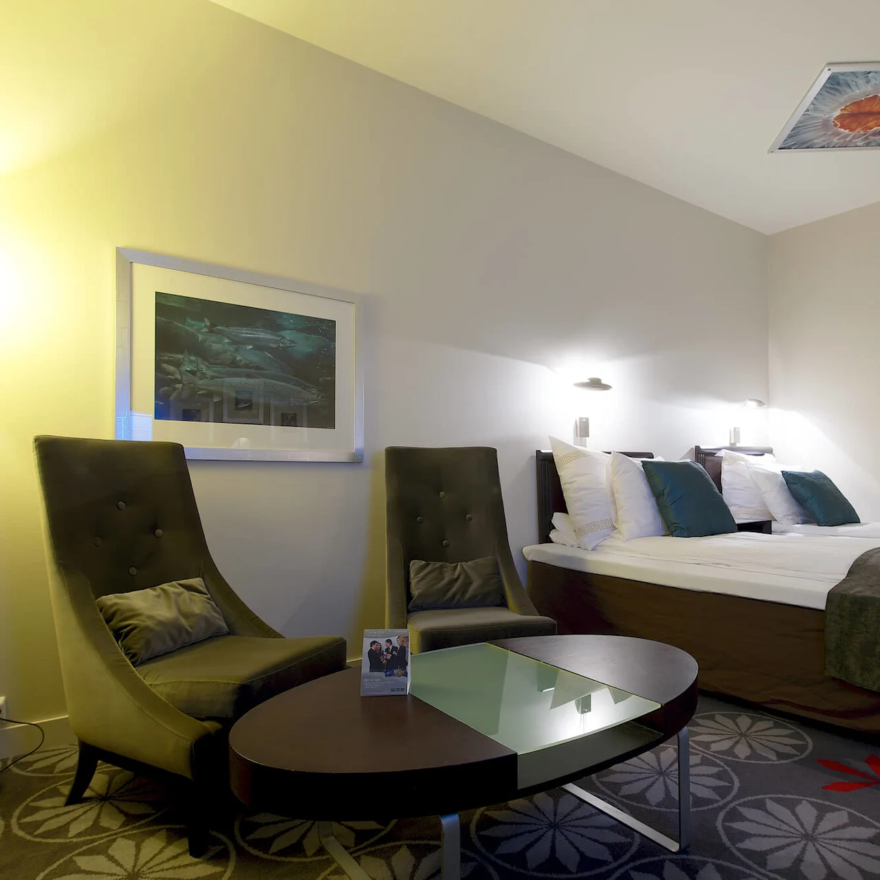 A well-lit hotel room with a double bed, two olive green armchairs, a floor lamp, a glass coffee table, and framed artwork on the walls.