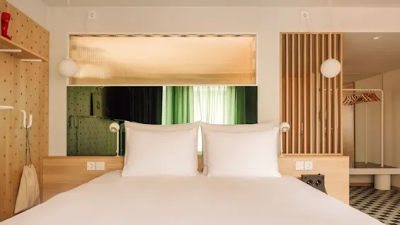 A contemporary hotel room at Hobo Oslo with a neatly made bed, light wood paneling, and a vibrant green curtain visible through a window.