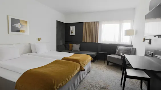 Two neatly made twin beds topped with mustard-yellow throws stand in a modern hotel room, contrasted by a gray sofa and armchair near a window with drawn white curtains.