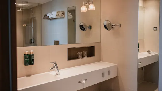 Sink with chrome faucet in a modern bathroom. Above, a large mirror with two wall lamps, toiletries, and towels. Shower area visible in the background.