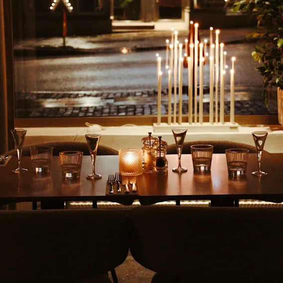 Warmly lit restaurant table set with wine glasses, cutlery, and a central candle, with a glowing electric advent candlestick in the window.