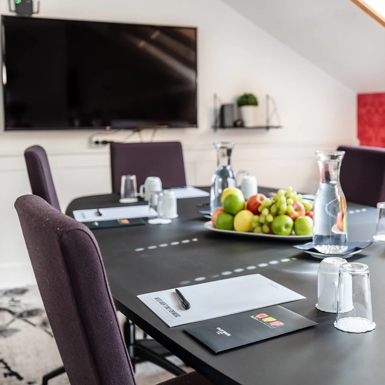 A modern conference room setup with notebook, pen, fruit platter, and water jug on a black table, under a large TV mounted on a white wall.