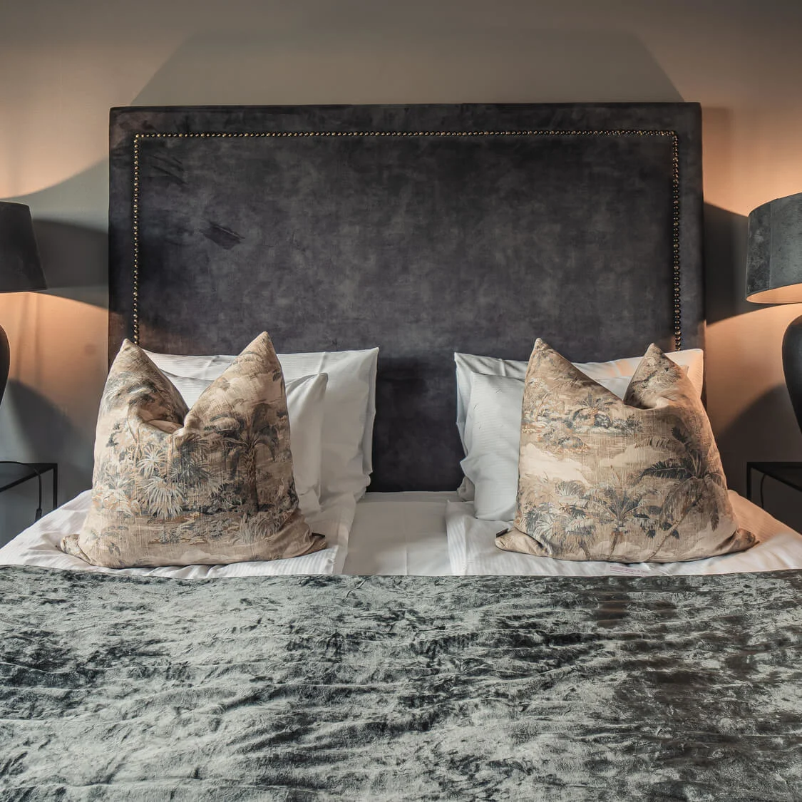 A bed with patterned pillows and a dark headboard is flanked by two nightstands with large black lamps, set in a softly lit bedroom.
