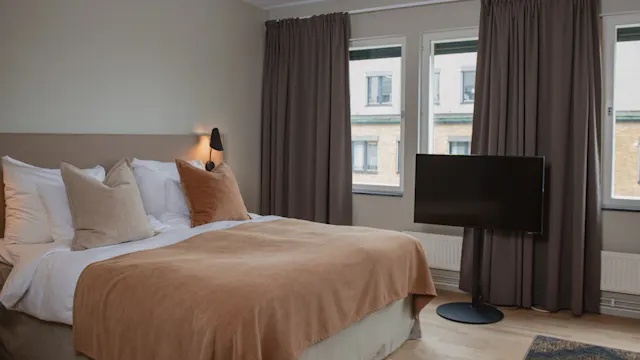 En säng med brunt sängkläder och kuddar står intill en vägglampa i ett rum med trägolv. En TV står motstående, nära gardinförsedda fönster.