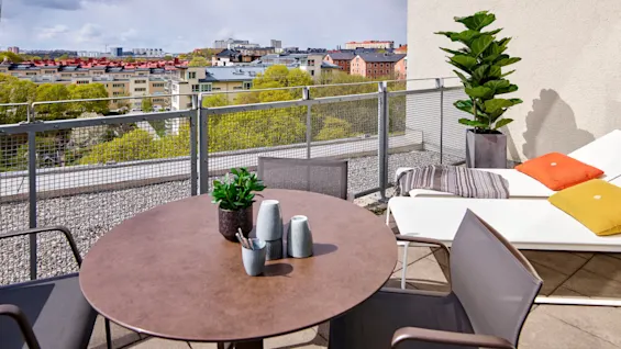 Et rundt bord med dekorative vaser står på en takterrasse, omgitt av stoler. Området har en pottet plante, solsenger og utsikt over byens bygninger og grøntområder.
