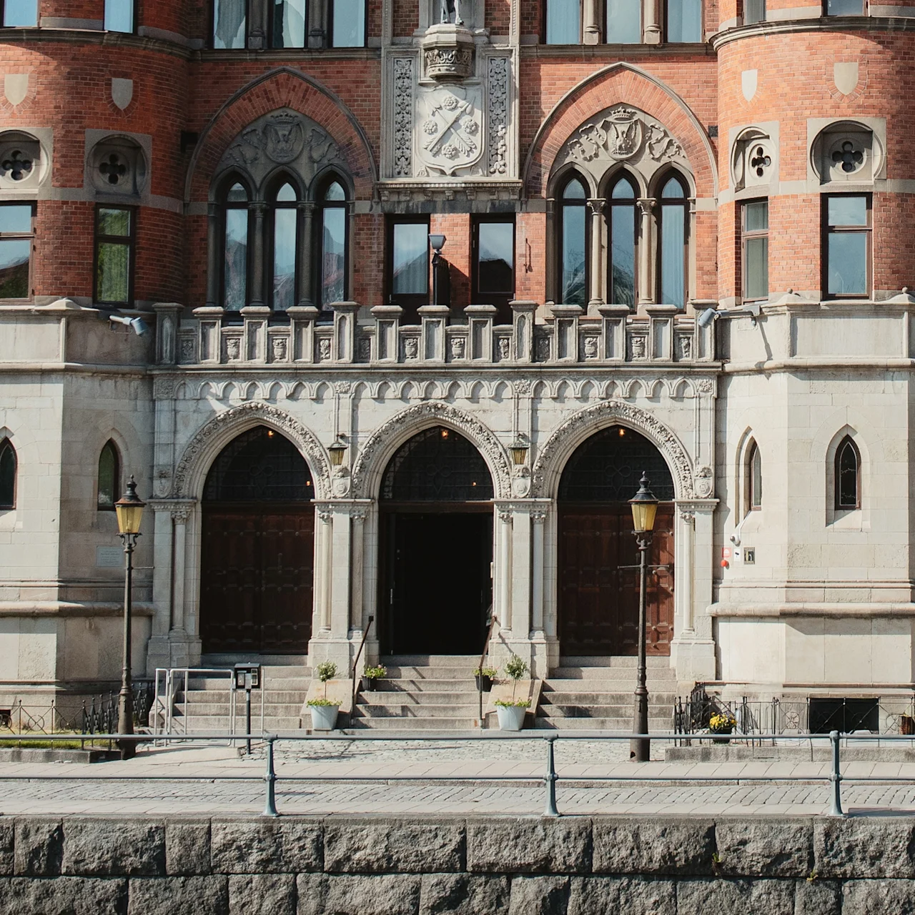 En historisk byggnadsfasad med tre välvda dörrar med intrikata stenutsmyckningar, omgiven av rött tegel och höga fönster. Text på en liten skylt lyder: "The Bishops Arms."
