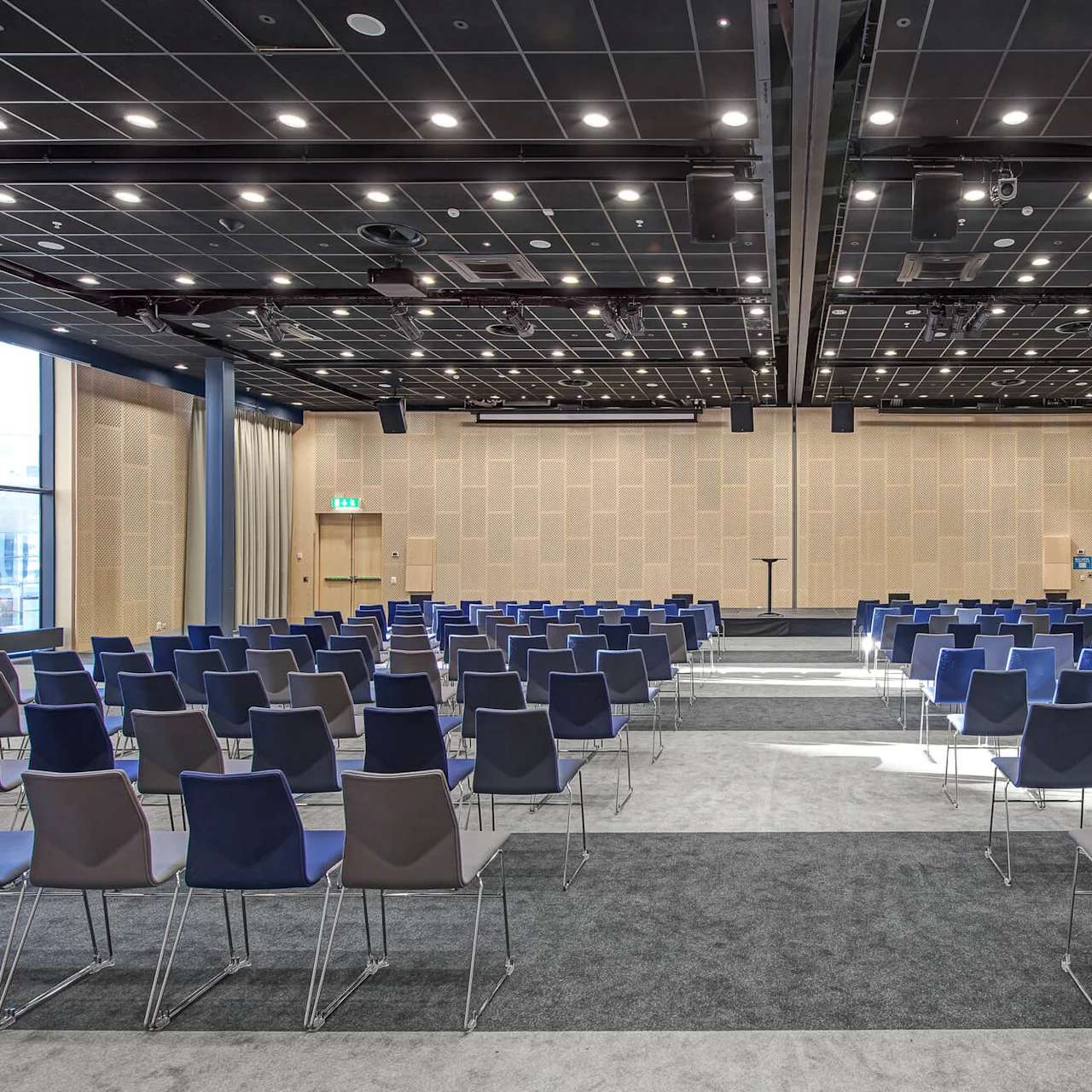 Rækker af tomme blå og grå stole vender mod en scene med et podium i et stort konferencelokale, under et sort gittermønstret loft med stærke lys.