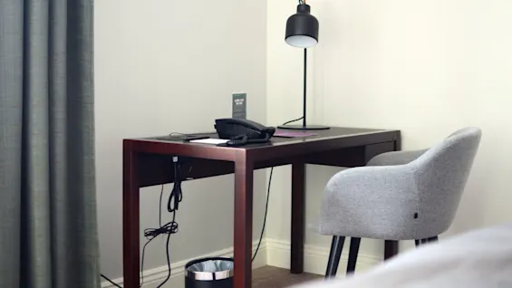 Et hotellskrivebord med en lampe, telefon, notisblokk og en stol, plassert nær et vindu med gardiner.