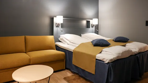 Säng med kuddar och filt, bredvid en senapsgul soffa, under två vägglampor; allt i ett modernt, mjukt upplyst rum med trägolv.