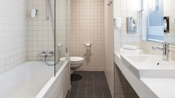 A modern bathroom features a bathtub with a glass screen, a wall-mounted shower, and a toilet. The space is tiled in white, with a sink and mirror to the right.