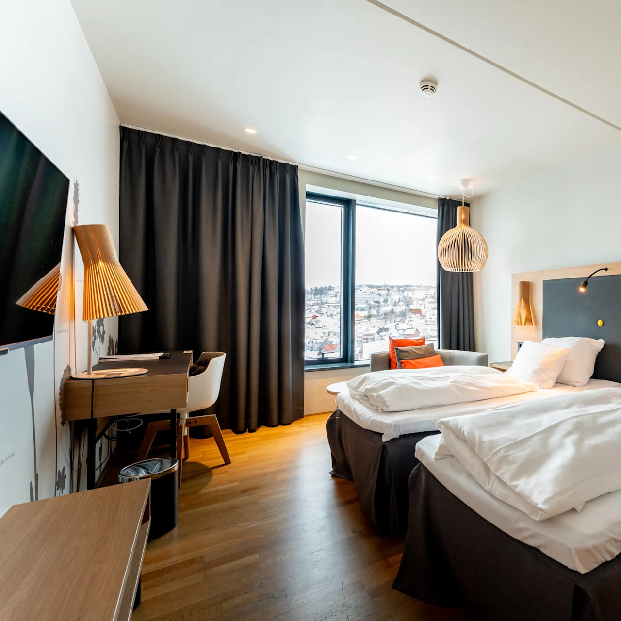 To velredte senge i et moderne hotelværelse, indrammet af trædetaljer og varm belysning, med et stort vindue viser et snedækket bylandskab.