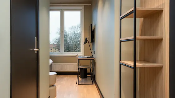 En mysig rumsentré med skrivbord, stol och hylla, som leder till ett ljust fönster med utsikt över träd.