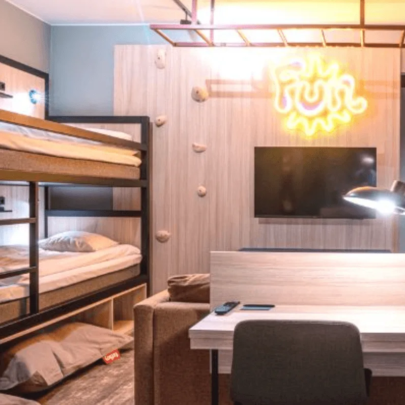 Bunk beds against a wall with climbing holds. A neon sign reads "Fun." A nearby desk, chair, and television create a cozy room setting.