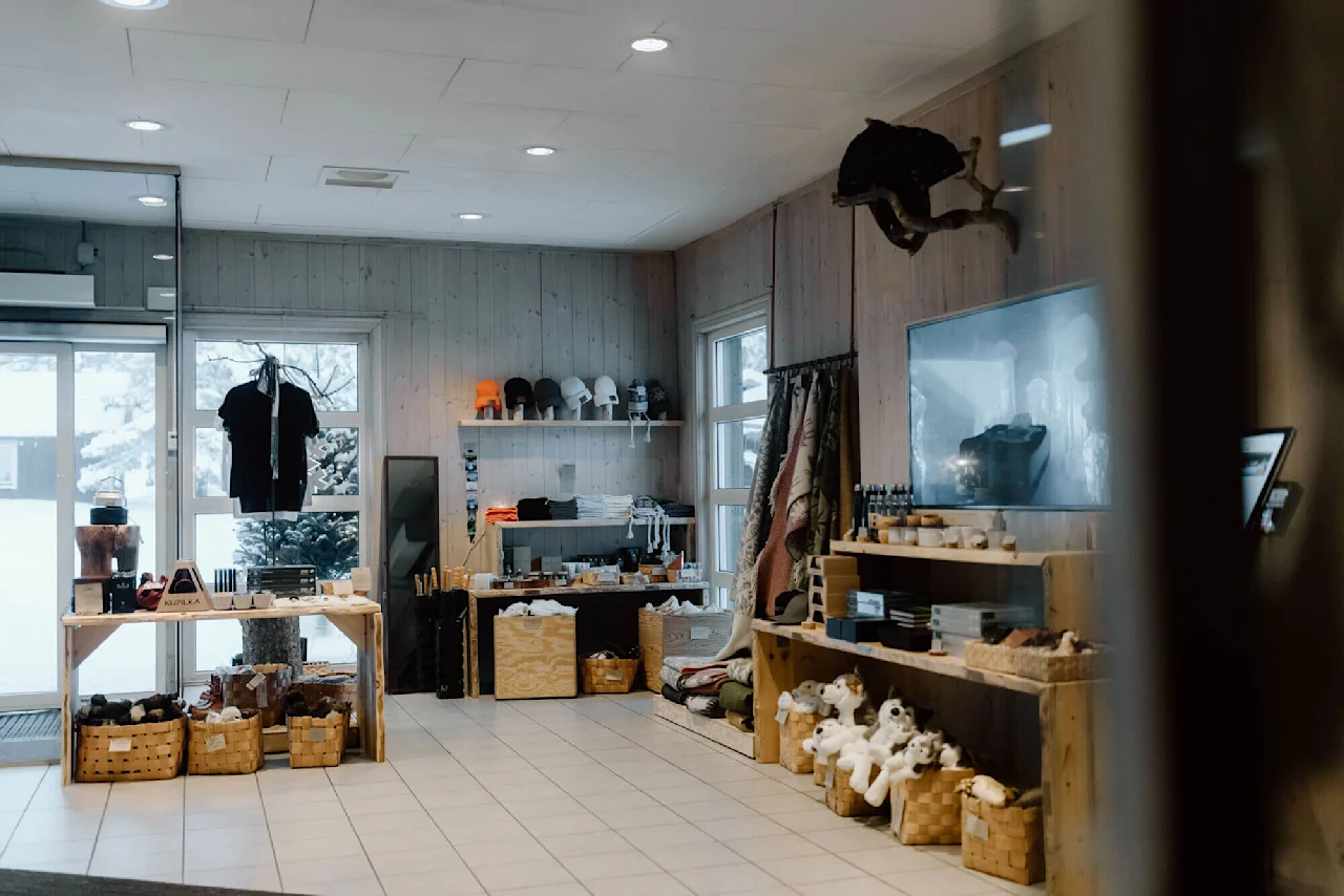 En butik viser forskellige ting som tøjdyr, kurve og tøj i en træindrettet, godt belyst indretning med snedækket udendørs udsigt gennem vinduerne.