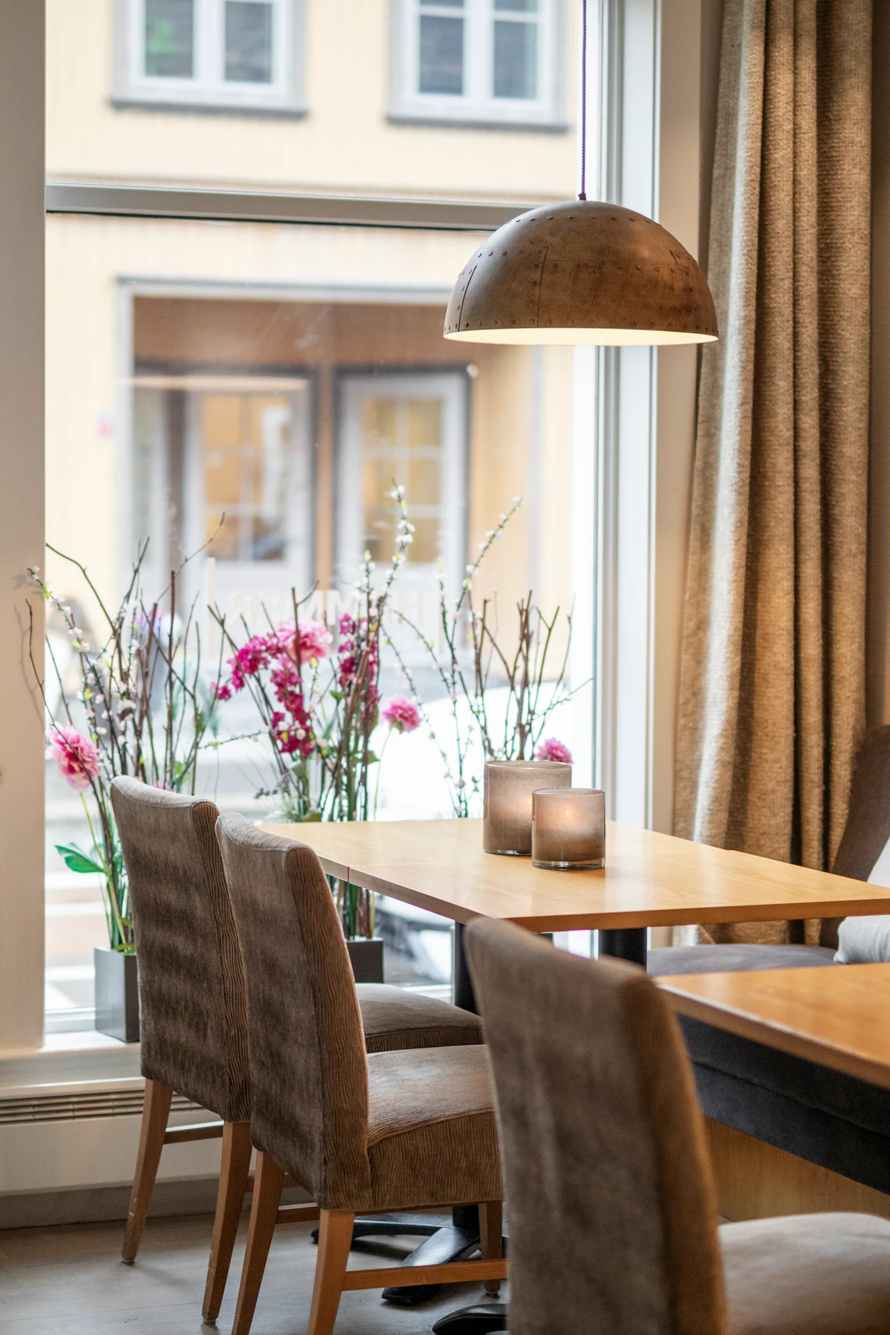 Et træbord omgivet af polstrede stole, med to stearinlys, står under en hængende lampe. Lyserøde blomster i vaser står ved vinduet, med udsigt over en gade.
