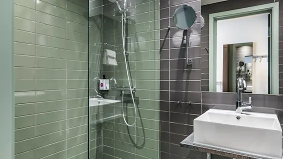 A modern bathroom features a glass-enclosed shower with green and gray tiles, a square white sink, and a round mirror on the wall, reflecting the surroundings.