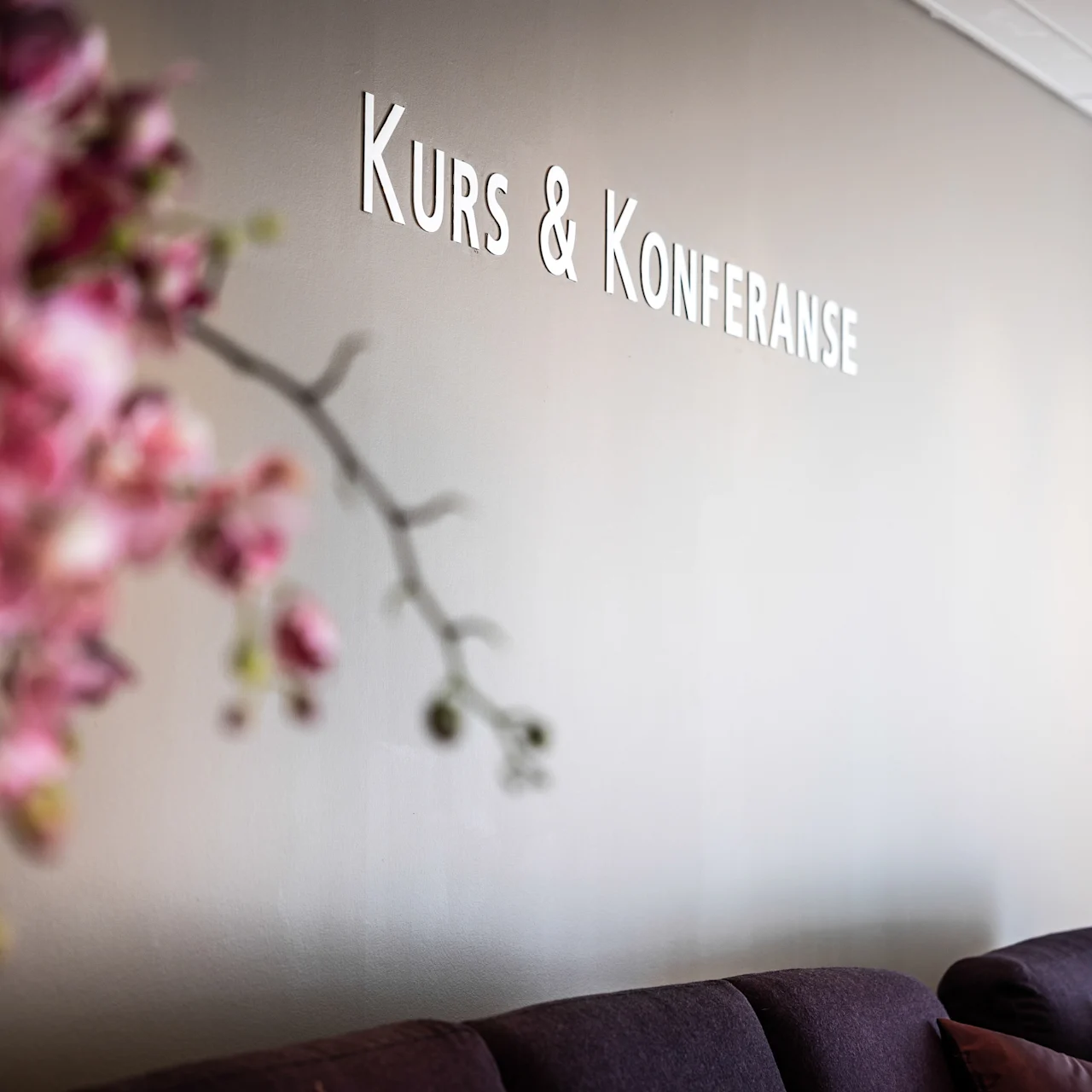En suddig rosa blomsterarrangemang i förgrunden med en väggskylt där det står "KURS & KONFERANSE" i bakgrunden, ovanför en mörk soffa.