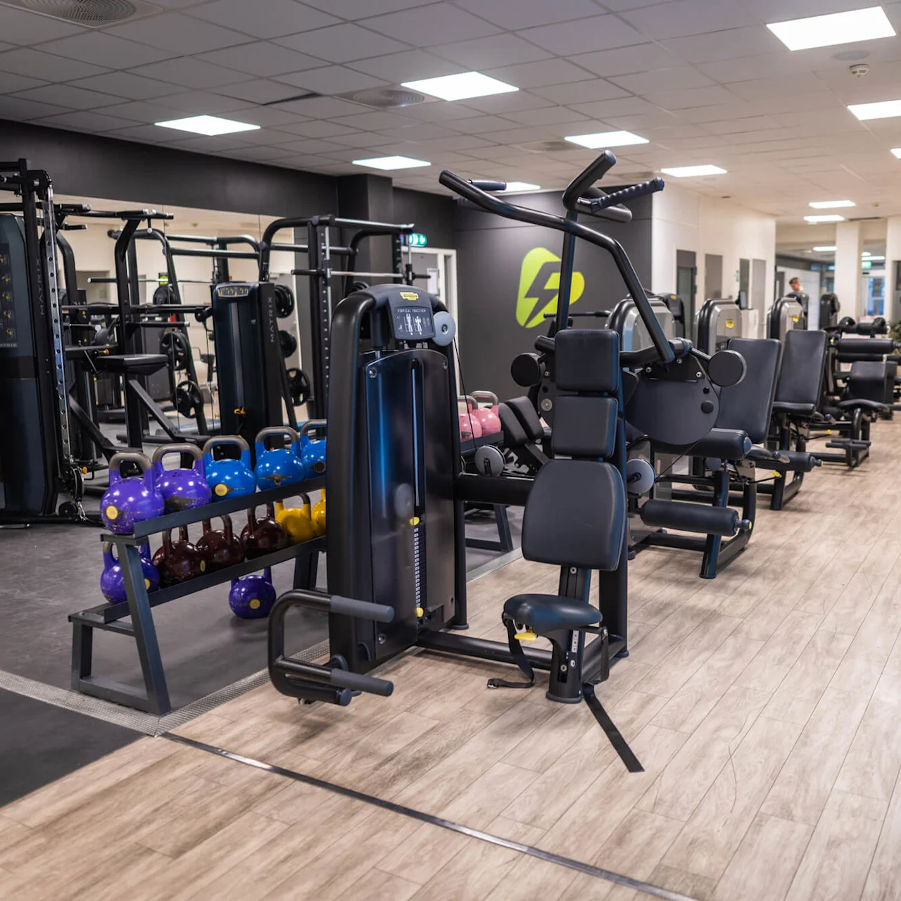 Træningsudstyr er pænt arrangeret i et godt oplyst fitnesscenter. Maskiner, kettlebells og en benpresser optager pladsen, omgivet af trægulv og hvidt fliset loft.