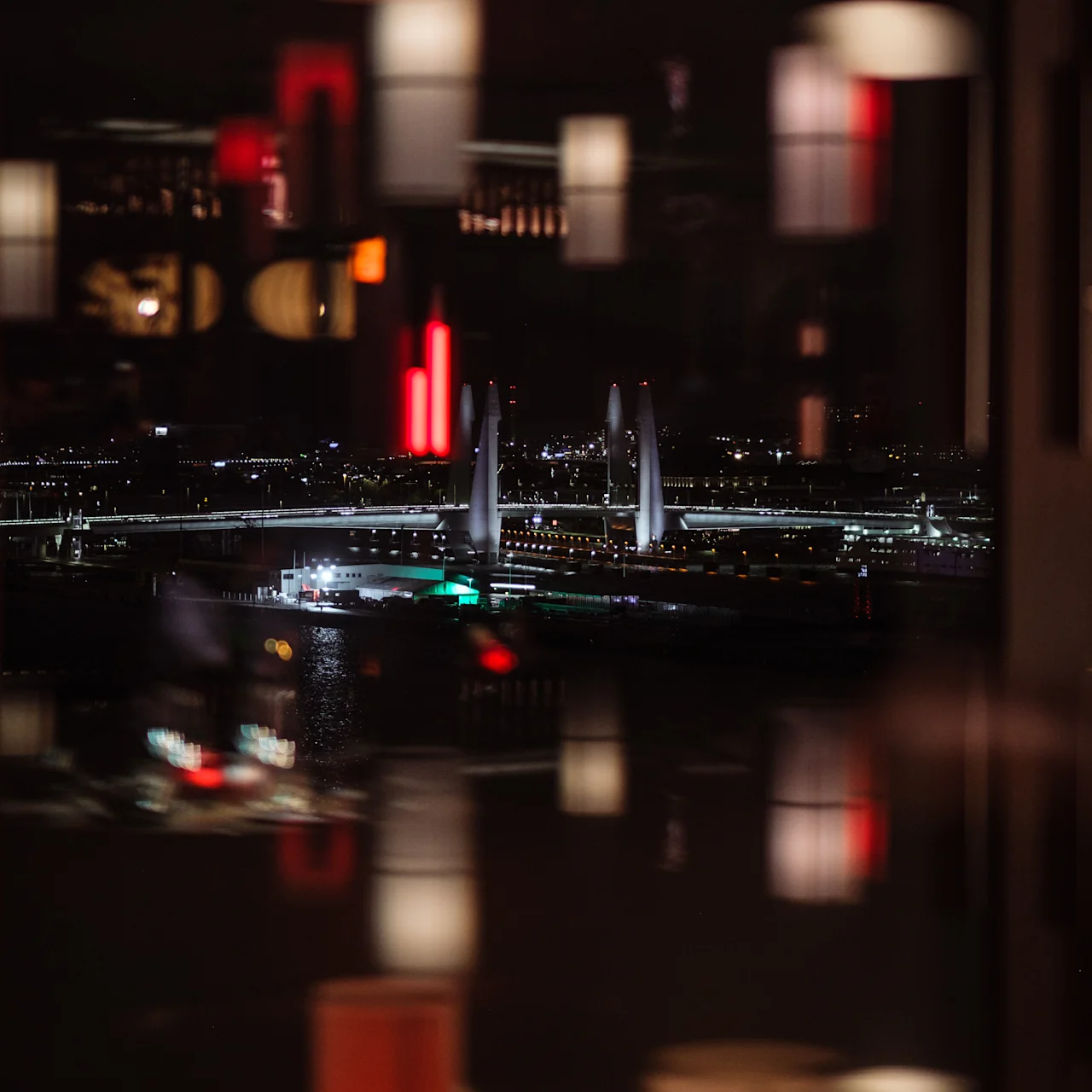 Röda och vita hängande lampor reflekteras i glas; en ljust upplyst bro sträcker sig över en stadsbild, som lyser upp natten med utspridda ljus.