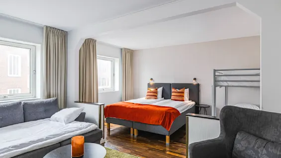 Et koselig hotellrom med en dobbeltseng kledd i livlige oransje detaljer. En sofa står ved vinduet, og en køyeseng i hjørnet. Myk belysning og varme toner skaper en innbydende atmosfære.