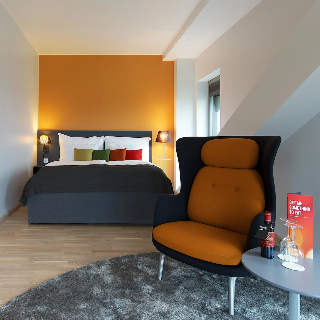 Ett modernt hotellrum med en prydligt bäddad säng med färgglada kuddar mot en orange vägg. En orange stol och ett litet bord med en vinflaska och glas. Text på kort: "GET ME SOMETHING TO EAT"