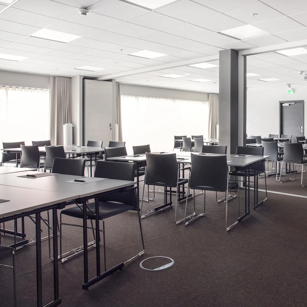 Konferensrum med tomma bord och stolar arrangerade i rader. Rummet är väl upplyst med stora fönster, en projektor i taket och ett uppfriskningsbord med växter.