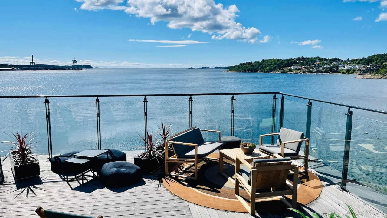 An outdoor patio with modern furniture overlooks a sparkling sea under a clear blue sky.