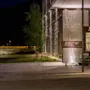Et skilt med teksten "SKIFER HOTEL" er plassert ved en opplyst inngang av en moderne bygning om natten, omringet av trær og en brosteinlagt sti. En bekk er synlig med en fjern bro i nærheten.