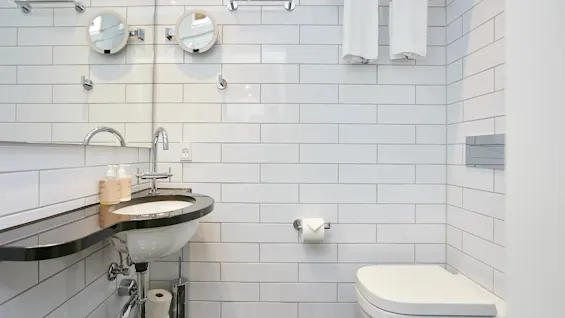Et rent, moderne badeværelse med hvide fliser, med håndvask, spejl, toilet og friske håndklæder.