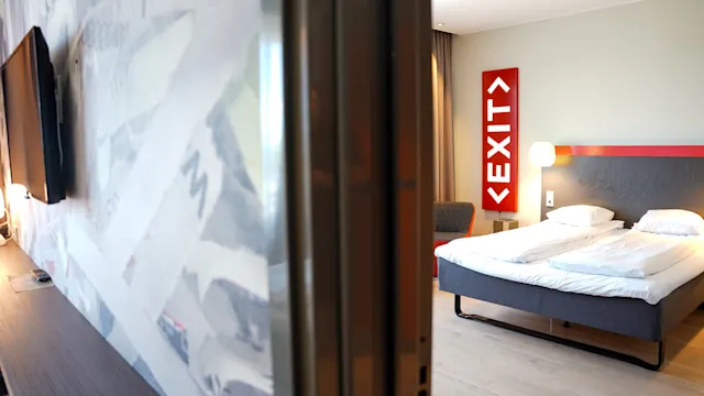Ett hotellrum har en prydligt bäddad säng på trägolv, med en röd "EXIT"-skylt på väggen bredvid. En TV är monterad på motsatt vägg.
