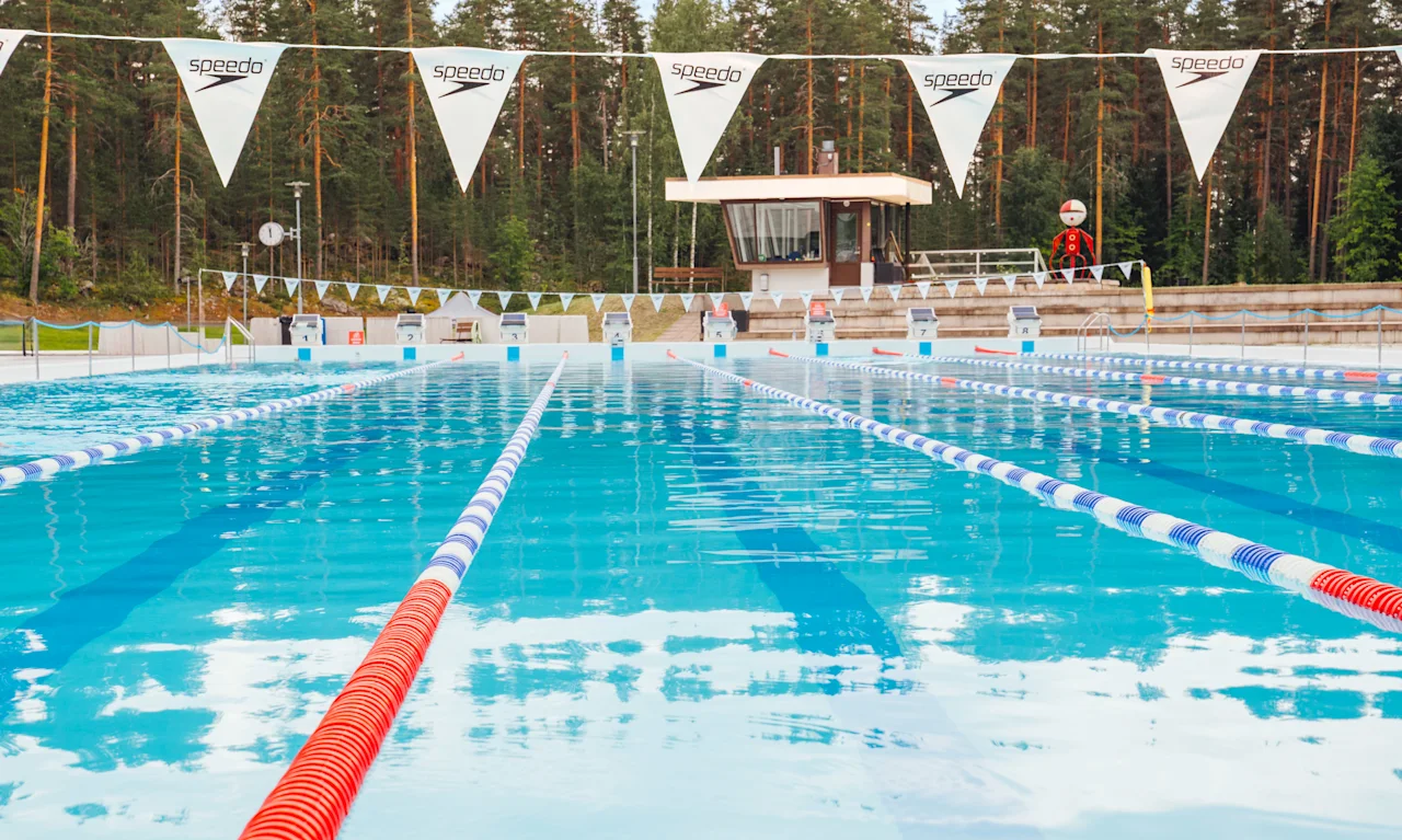 Ulkouima-allas selkeine ratoineen, ympärillä metsää taustalla. "Speedo" teksti liput roikkuvat altaan yllä. Uimavalvojan torni näkyvissä ja valvoja paikalla.