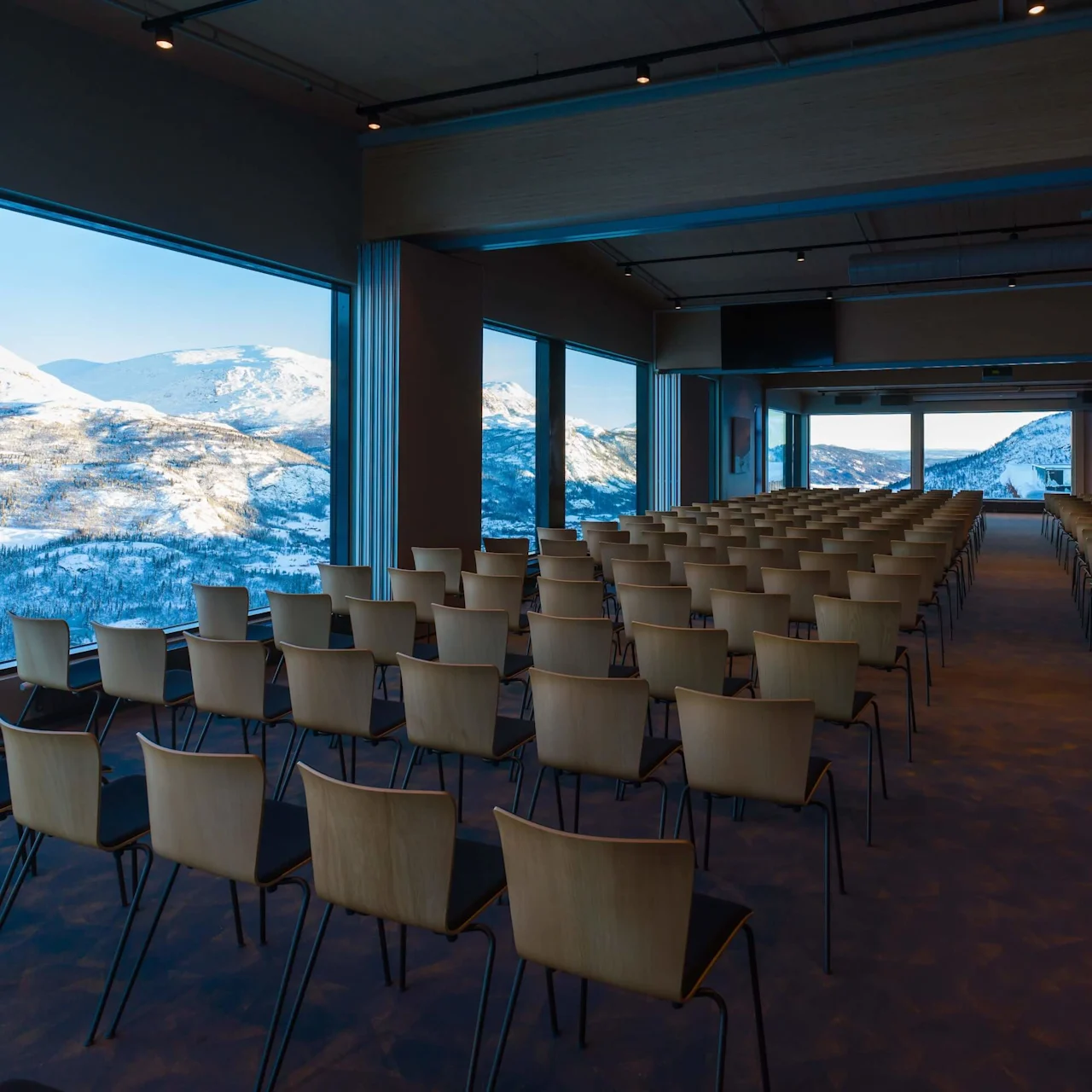 Rows of empty wooden chairs face forward in a spacious conference room with large windows showcasing a snowy mountain landscape outside.