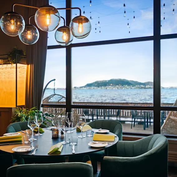 Spisebordet er elegant dekket med tallerkener og glass. Moderne sfæriske lys henger over. Stort vindu gir utsikt over kystlandskap og blå himmel med øy, som forbedrer den naturskjønne spiseomgivelsen.