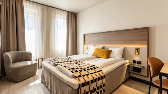 Viihtyisä hotellihuone, jossa mukava sänky, tyylikäs nojatuoli ja työpöytäalue, kylpee luonnonvalossa ikkunoista.