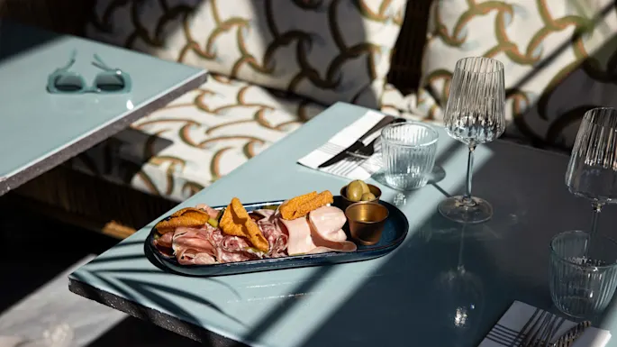 A charcuterie platter with cold cuts, crisps, and olives sits on a light blue table. Nearby, empty glasses and silverware are neatly arranged under dappled sunlight with patterned cushions.