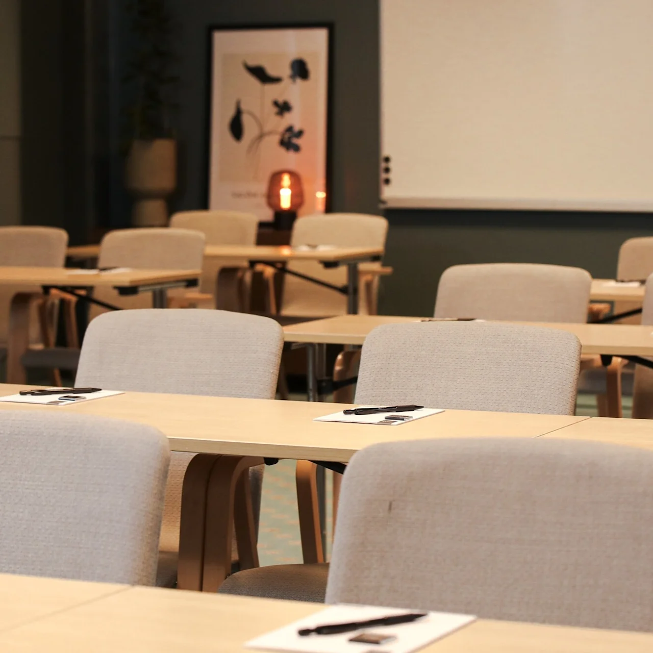 Rows of beige chairs and wooden desks, set up in a classroom, each with a notebook and pen. A soft light glows and a framed poster is visible in the background.