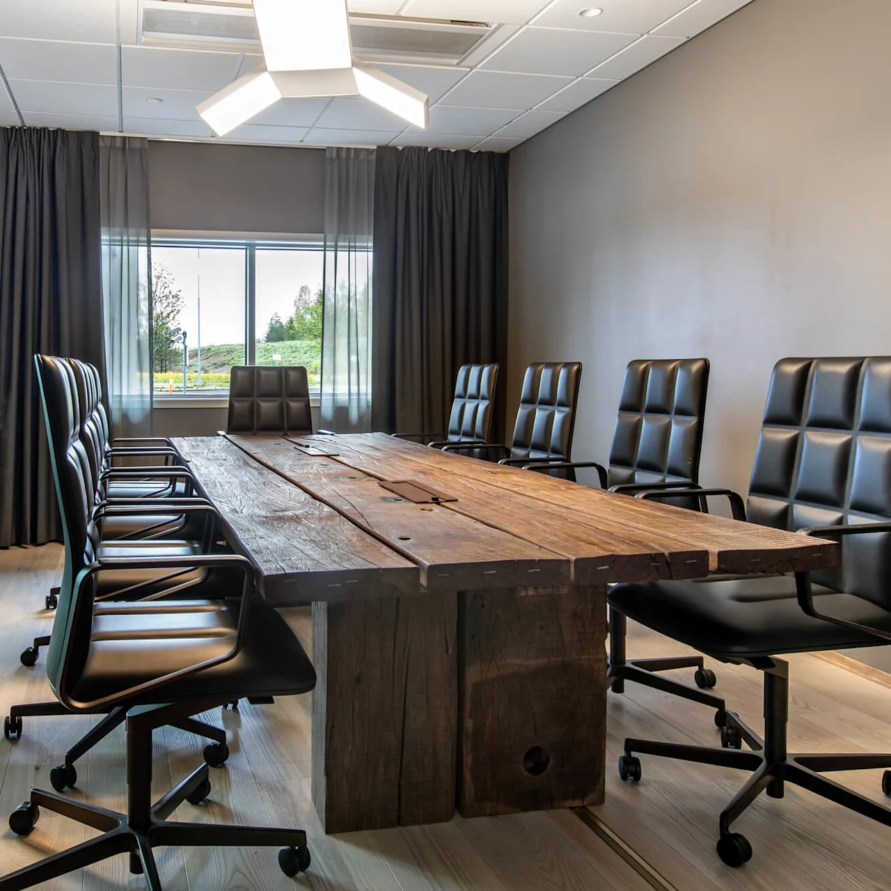 A large wooden conference table surrounded by black swivel chairs in a modern meeting room with a window view of greenery.