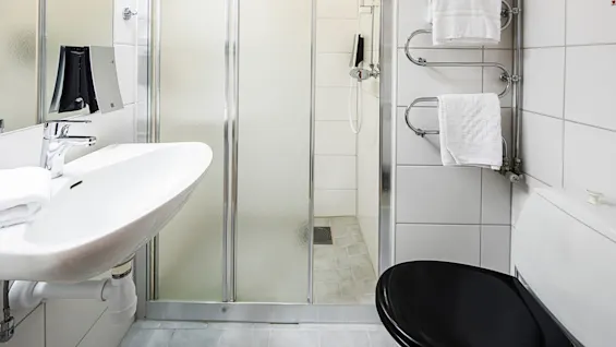 A white bathroom with a sink, mirror, shower, and black toilet bowl; towels hang on a rack.